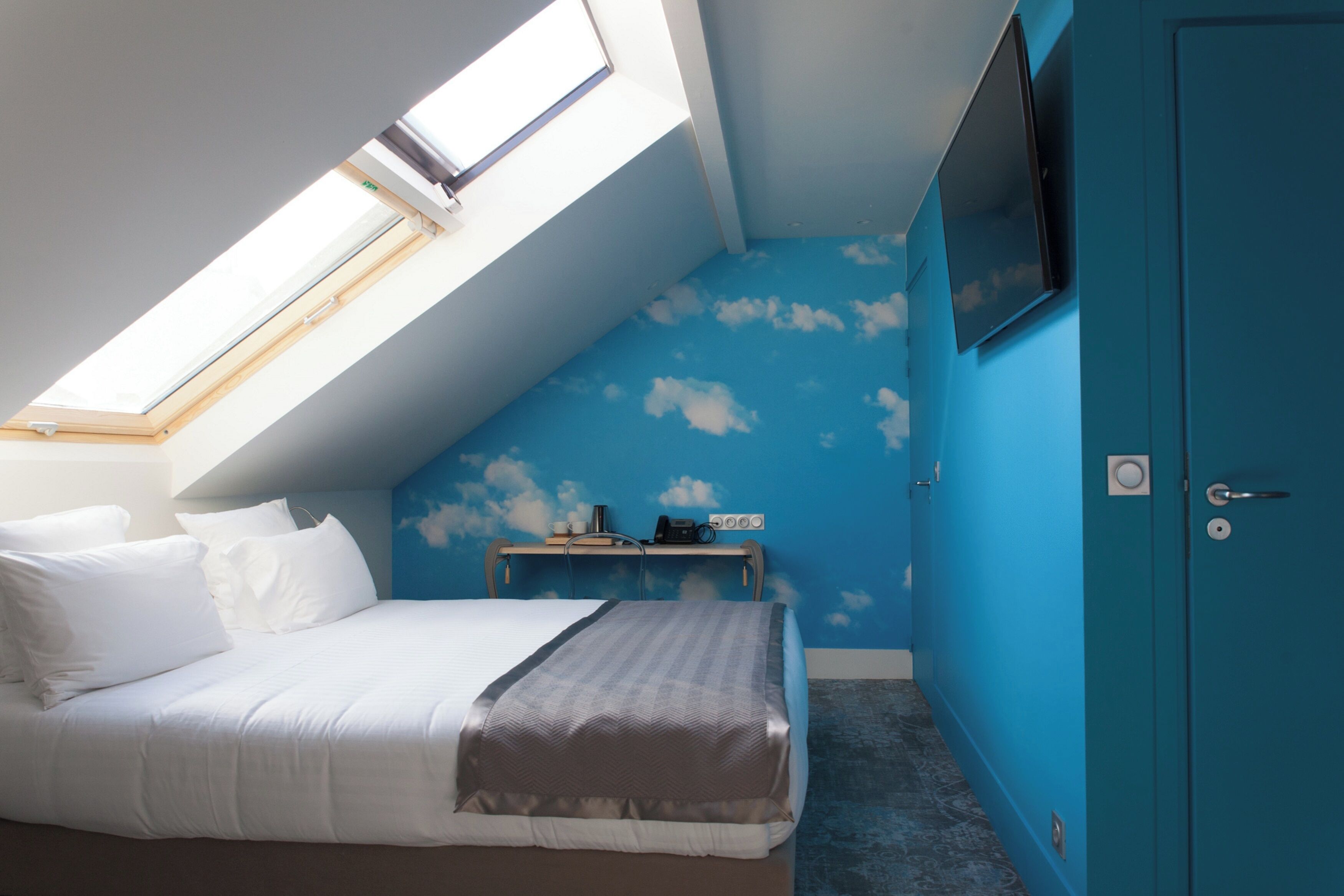 A bedroom with a skylight, a bed with white bedding, a blue wall with cloud patterns, a TV mounted on the wall, and a blue wardrobe.