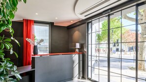 A reception area with a curved counter, a red curtain, a potted plant, and a view of the street outside. - Hotel Sixteen Paris Montrouge (Montrouge)