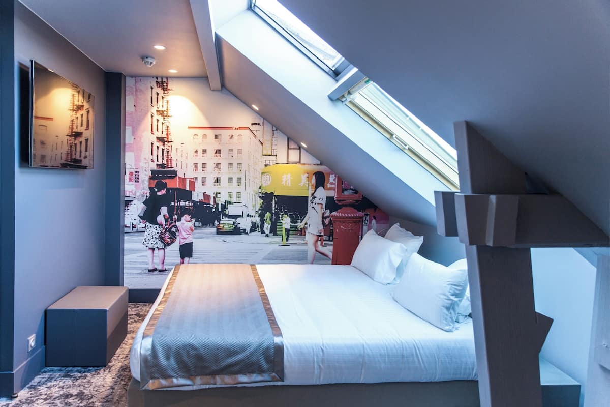 A modern hotel room with a large bed, a skylight, and a cityscape mural on the wall.