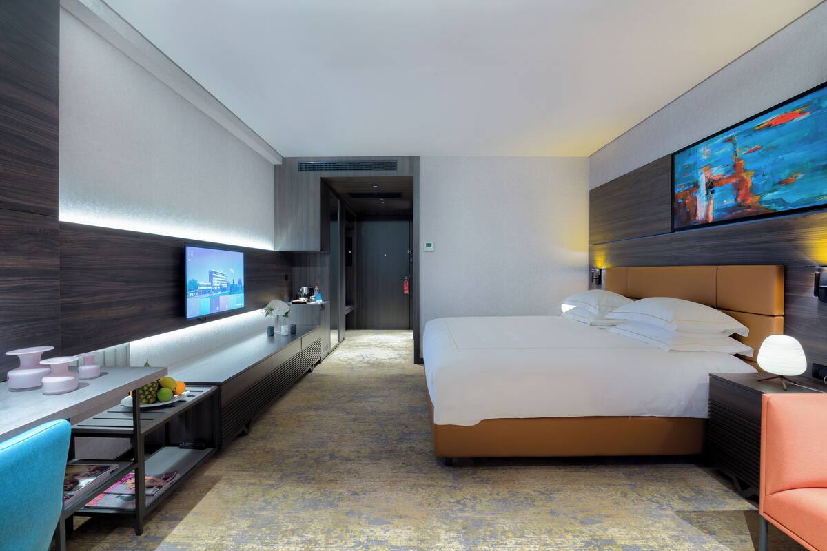 A modern hotel room with a large bed, a desk with a flat-screen TV, a seating area with a chair, and a painting on the wall.