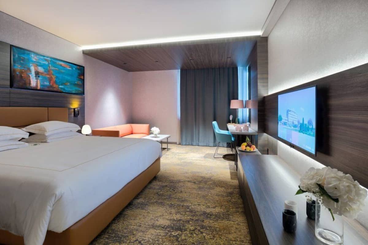 A hotel room with a large bed, a desk with a chair, a TV mounted on the wall, and a painting above the bed.
