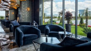 A modern lounge area with dark blue sofas and a large window offering a view of a landscaped garden and buildings. - Emerald Hotel Pristina, Kosovo (Preoce)