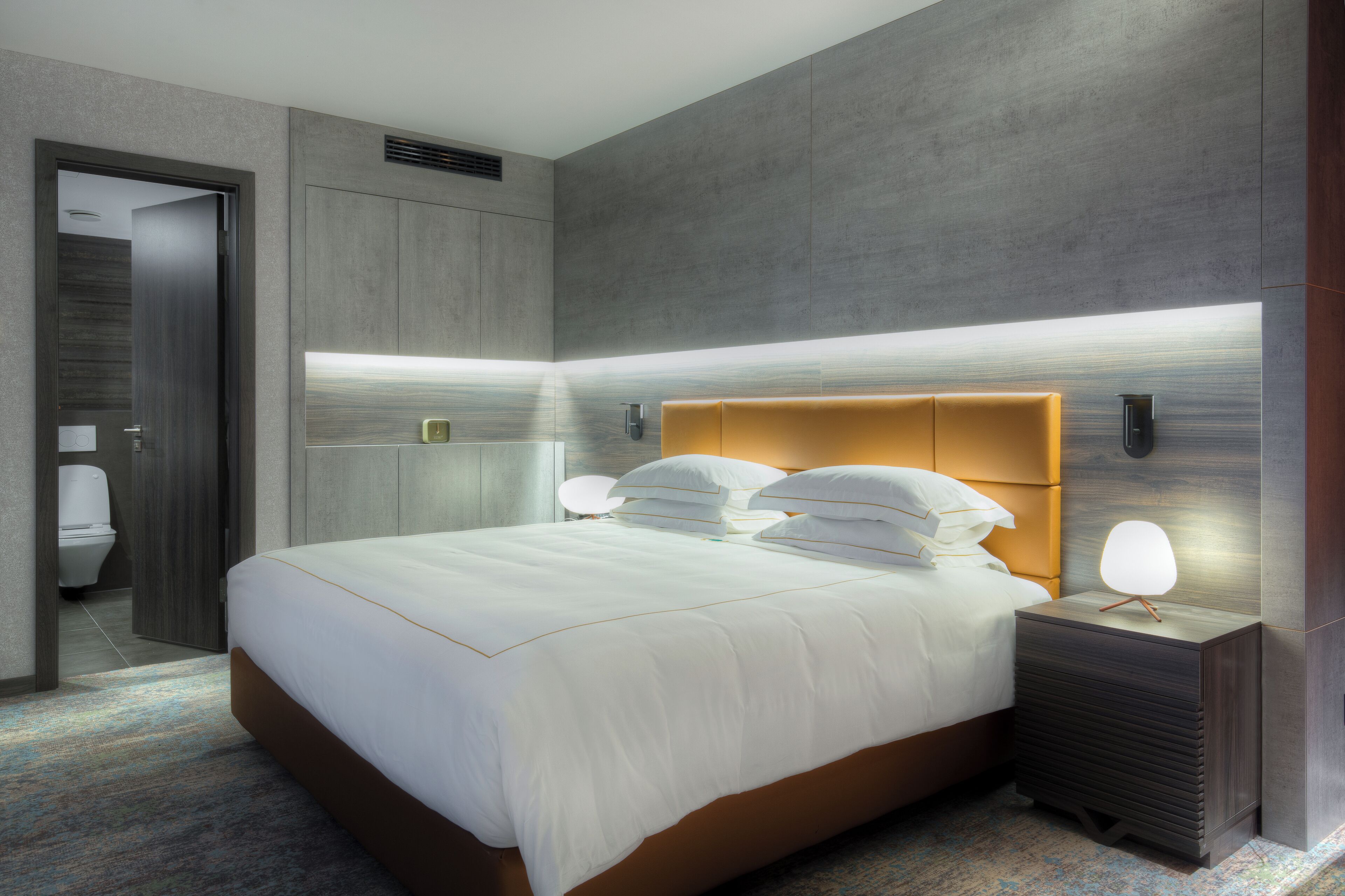 A modern hotel room with a large bed, bedside tables, and a bathroom visible through an open door.