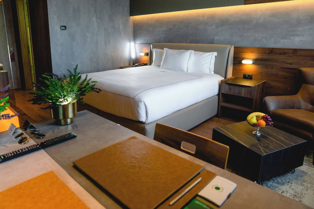 A modern hotel room with a large bed, bedside tables, a desk with a chair, a potted plant, and a fruit bowl.