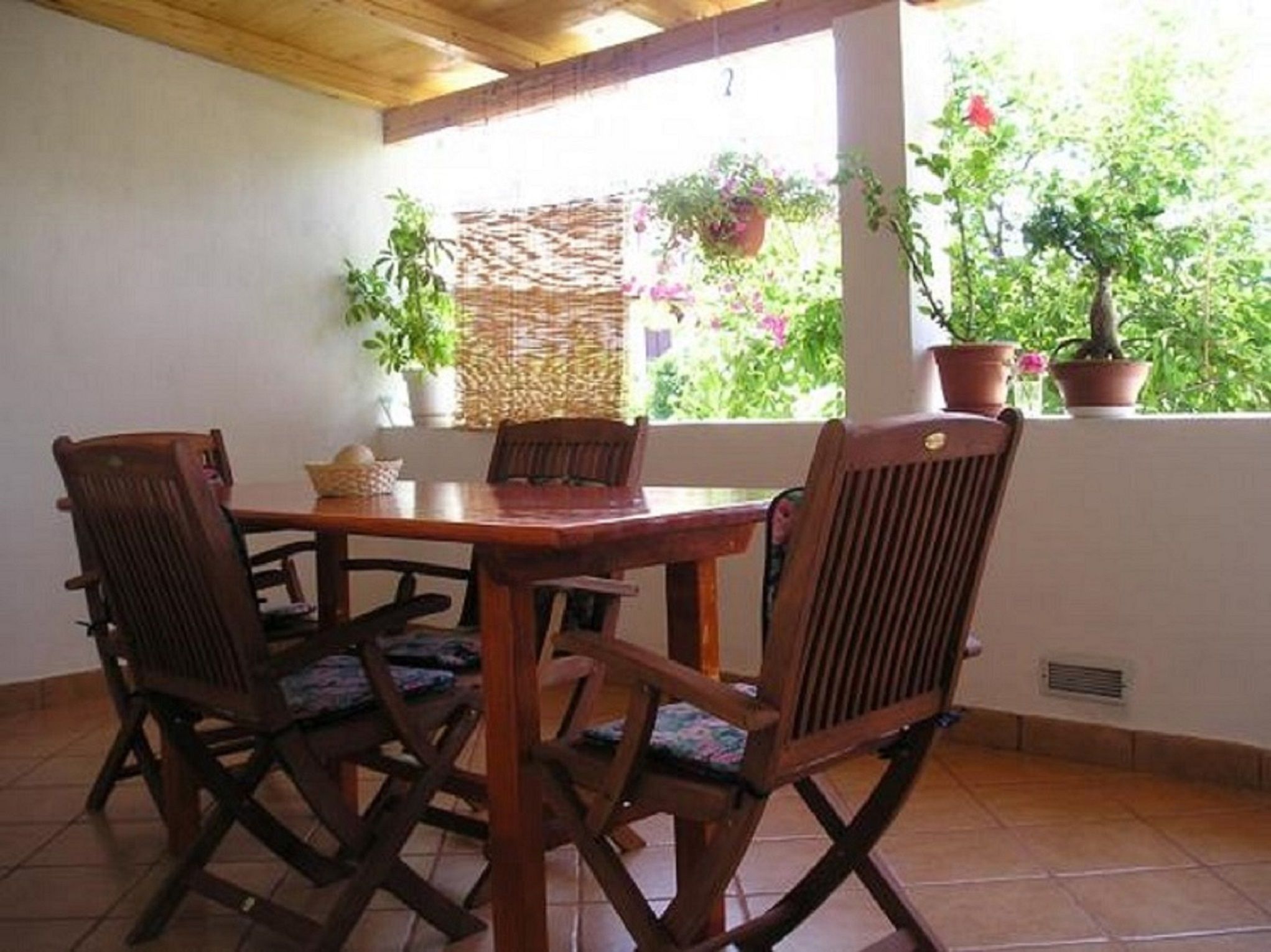 A covered patio with a wooden table and chairs, surrounded by potted plants and a view of greenery.