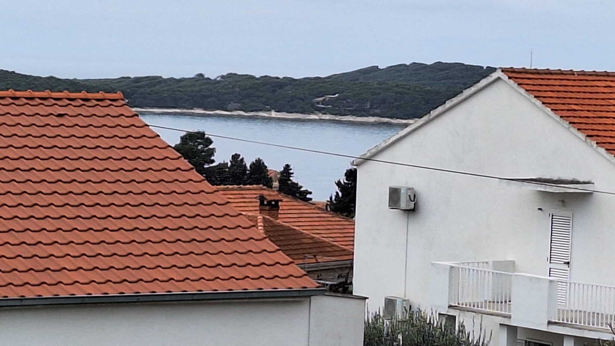 A coastal building with a red-tiled roof, a balcony, and a view of the sea and hills in the background.