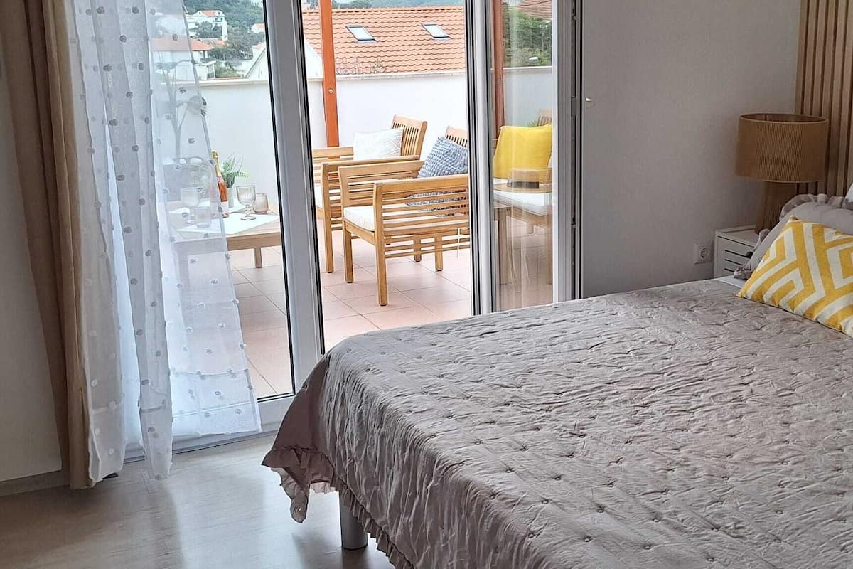 Ein Schlafzimmer mit Bett, Nachttisch, Lampe und Blick auf eine Terrasse mit Tisch und Stühlen.