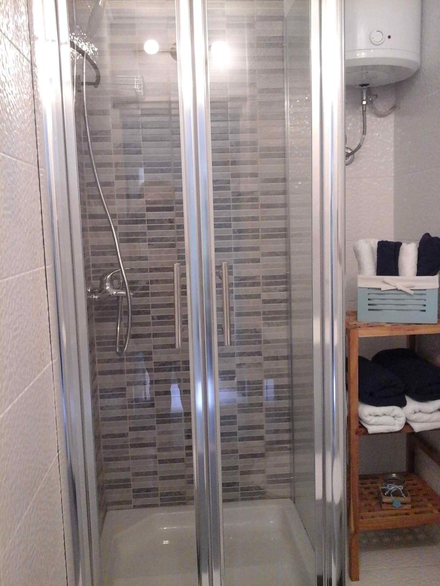 A glass-enclosed shower with a tiled wall, a handheld showerhead, and a control panel above the shower.