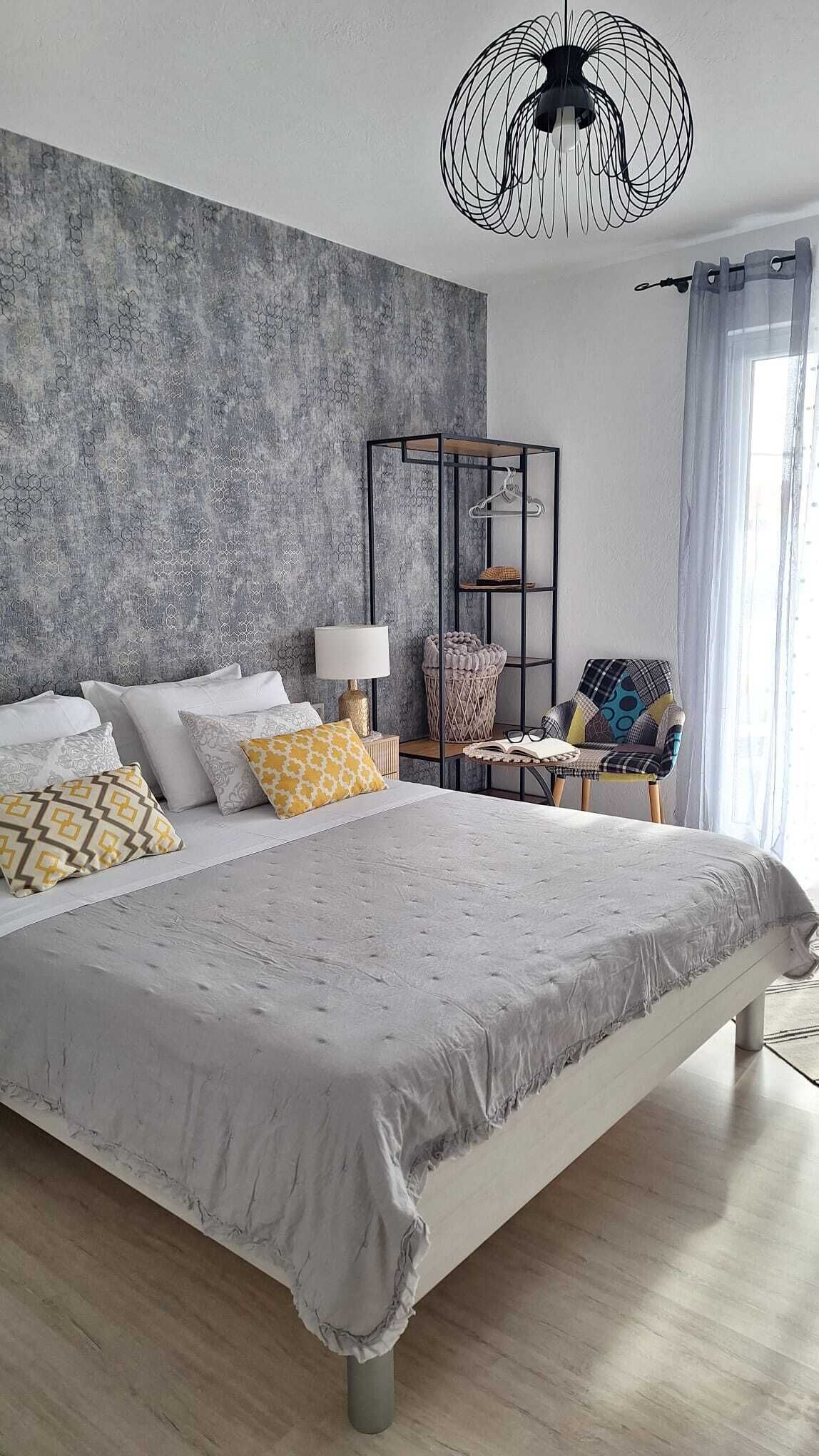 A modern bedroom with a large bed, a hanging light fixture, a side table with a lamp, a chair, and a decorative wall panel.