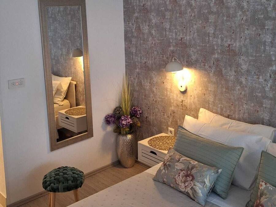 A bedroom with a large bed, a wall-mounted mirror, a textured wall, a floor lamp, and a bedside table with a vase of flowers.