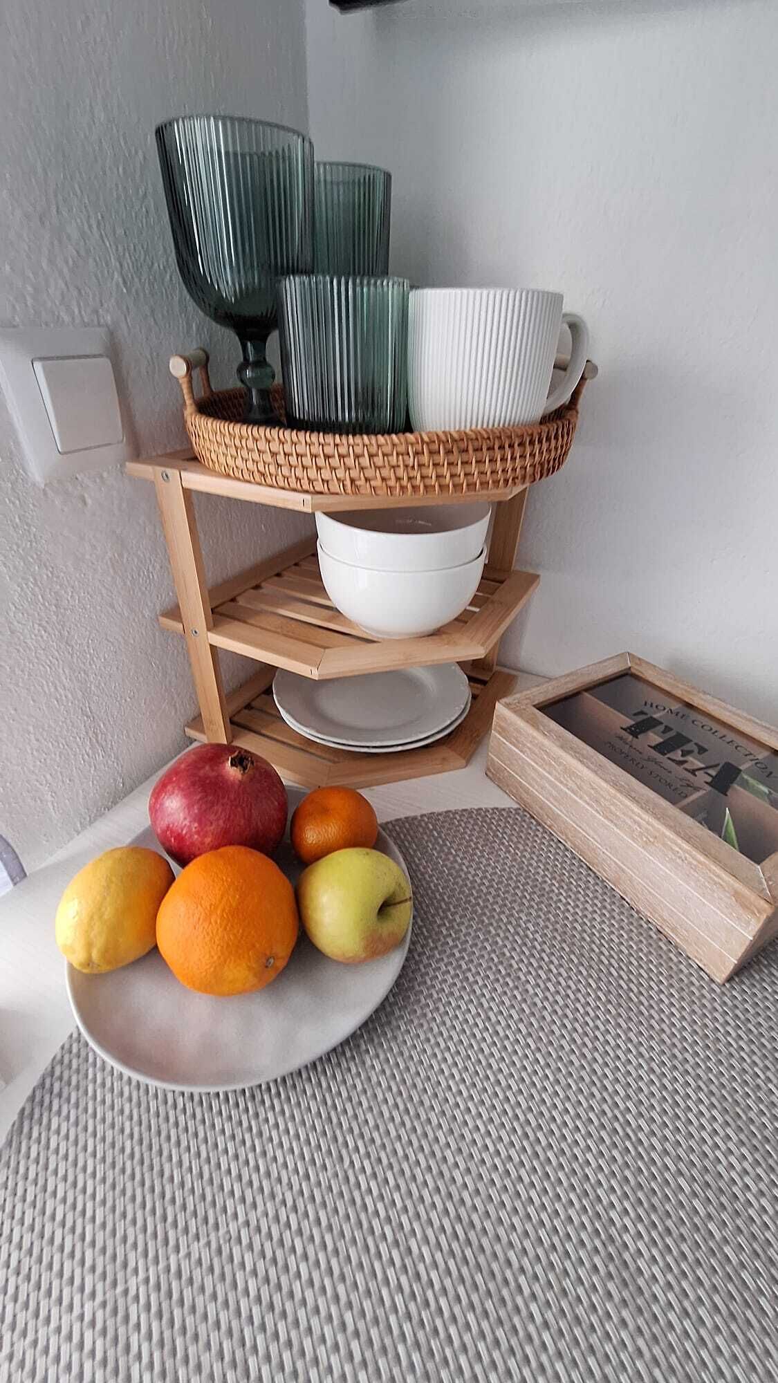 Eine Küchenzeile mit einer Obstschale mit Äpfeln, Orangen und Zitronen, einem Holzbodenregal mit Schüsseln und einem gerahmten Bild sowie einer strukturierten Matte auf dem Boden.