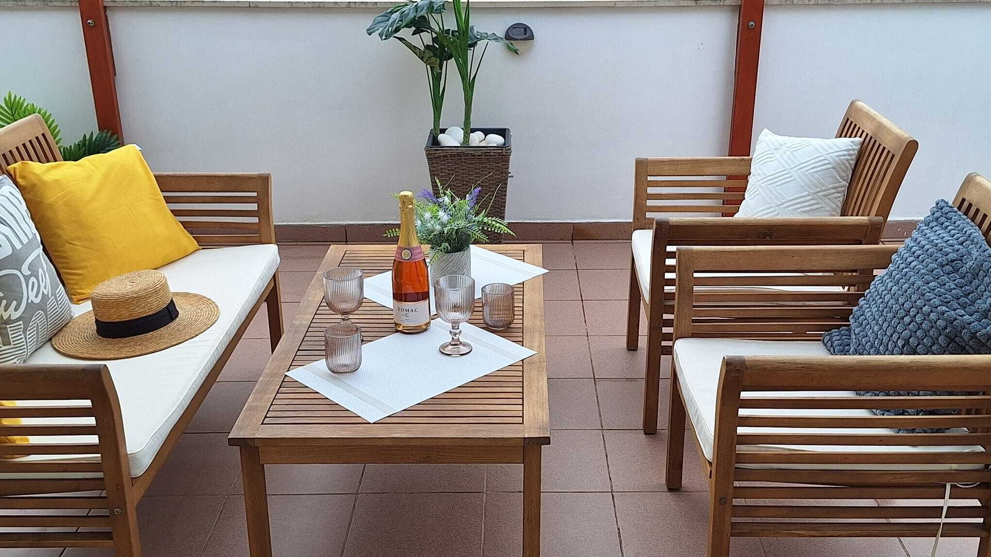 Un patio con muebles de madera, una mesa con una botella y vasos, y una planta en maceta.