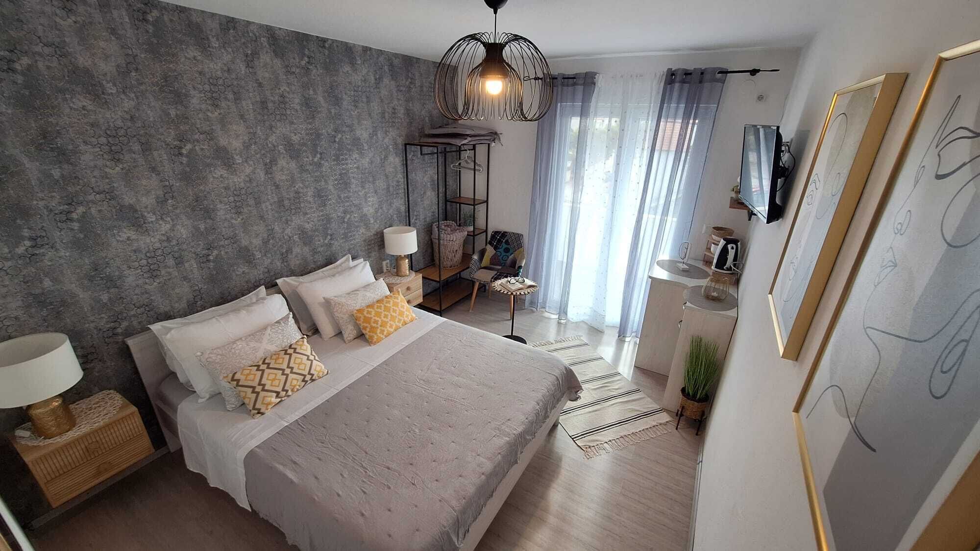 Un dormitorio moderno con una cama grande, una lámpara colgante, una mesita de noche pequeña, una silla, un televisor y una ventana con cortinas.