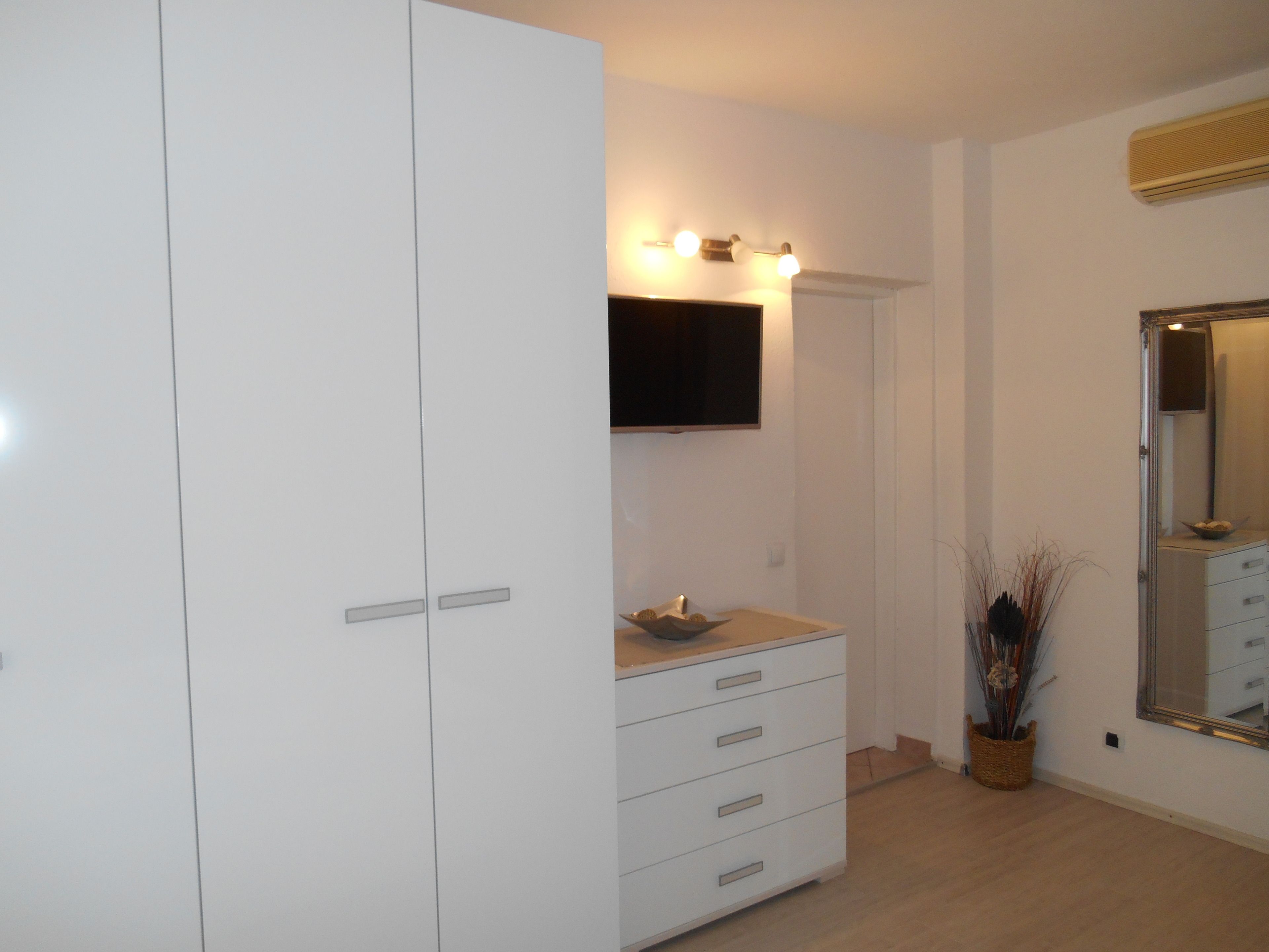 Un dormitorio con un armario blanco, una mesita de noche con lámpara, un televisor de pantalla plana montado en la pared y un espejo grande.