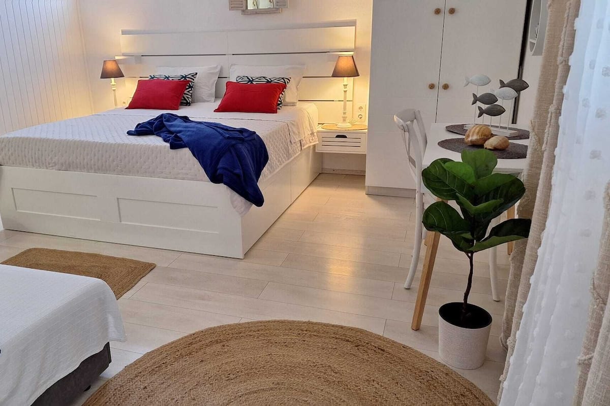 Ein modernes Schlafzimmer mit einem weißen Bett, Kissen mit roten und schwarzen Mustern, einer blauen Decke und einem Nachttisch mit einer Lampe. In der Ecke steht ein runder Holzschemel und ein Topf mit einer Pflanze.