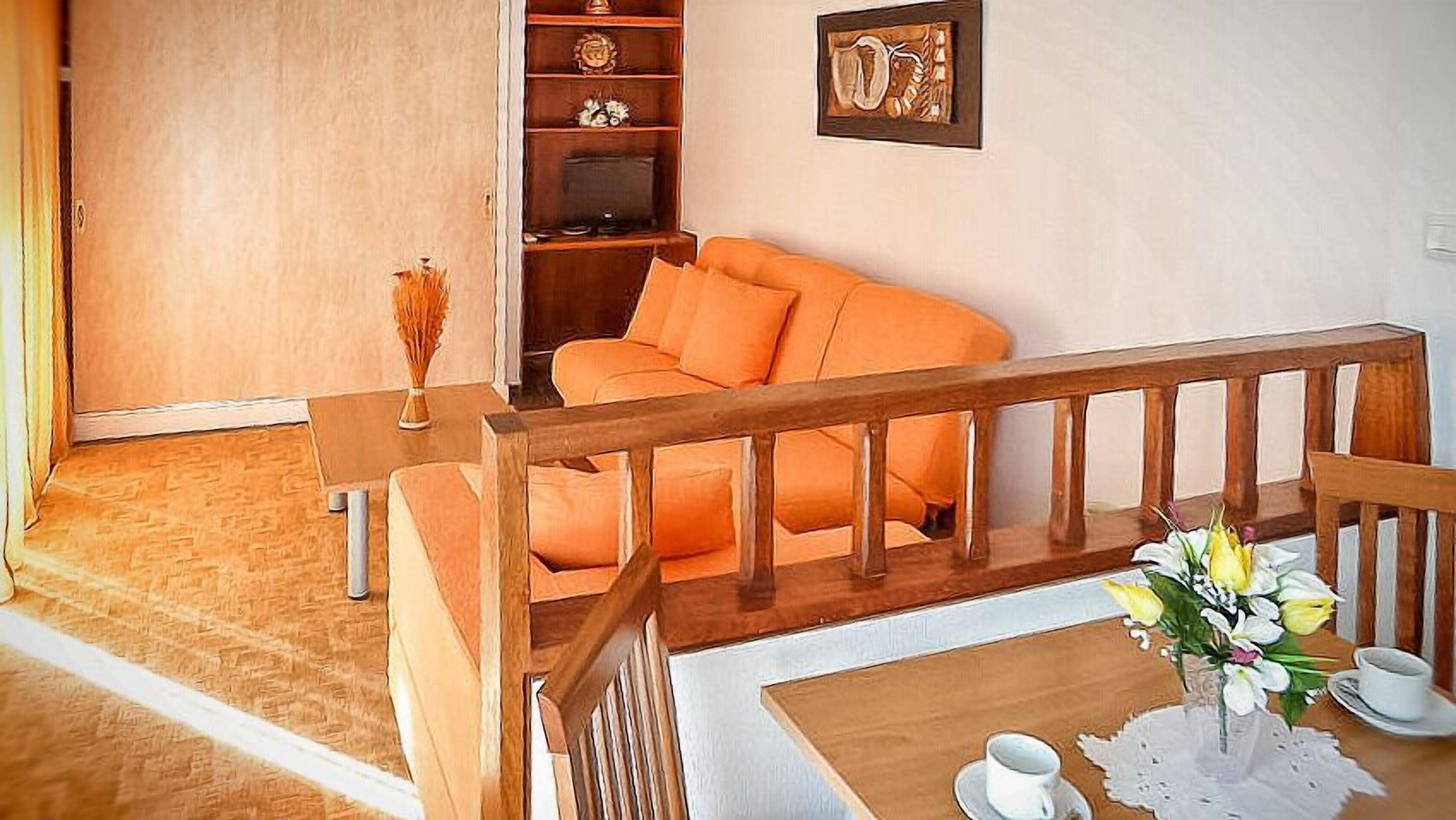 A cozy living room with a wooden sofa, a wooden coffee table with cups and a vase of flowers, and a bookshelf with decorative items.