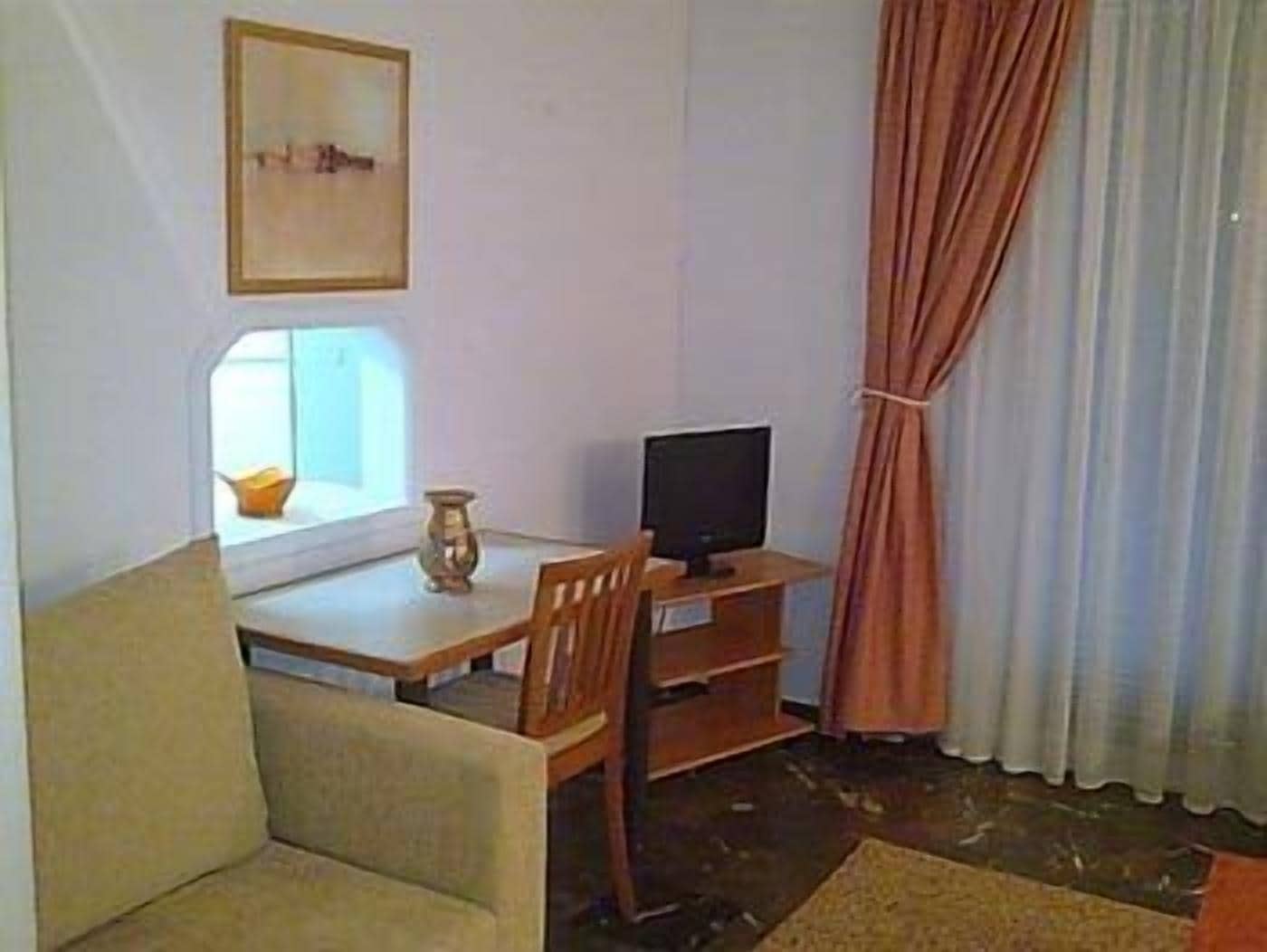 A small, furnished room with a wooden table, a television, a chair, and a window with curtains.
