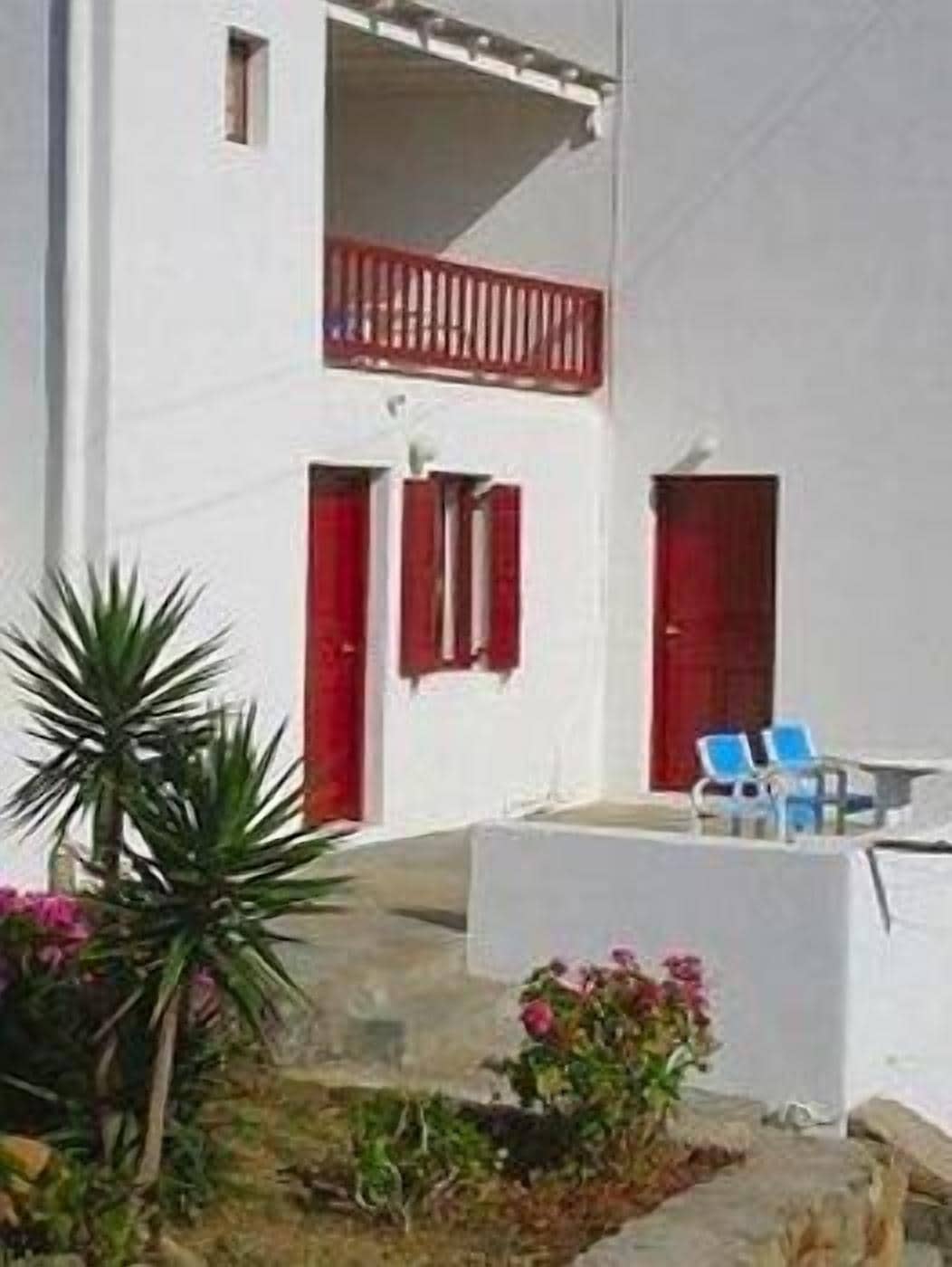 Un bâtiment blanc avec des portes et des fenêtres rouges, un balcon doté d’une balustrade rouge, et un petit jardin agrémenté de plantes et de fleurs.