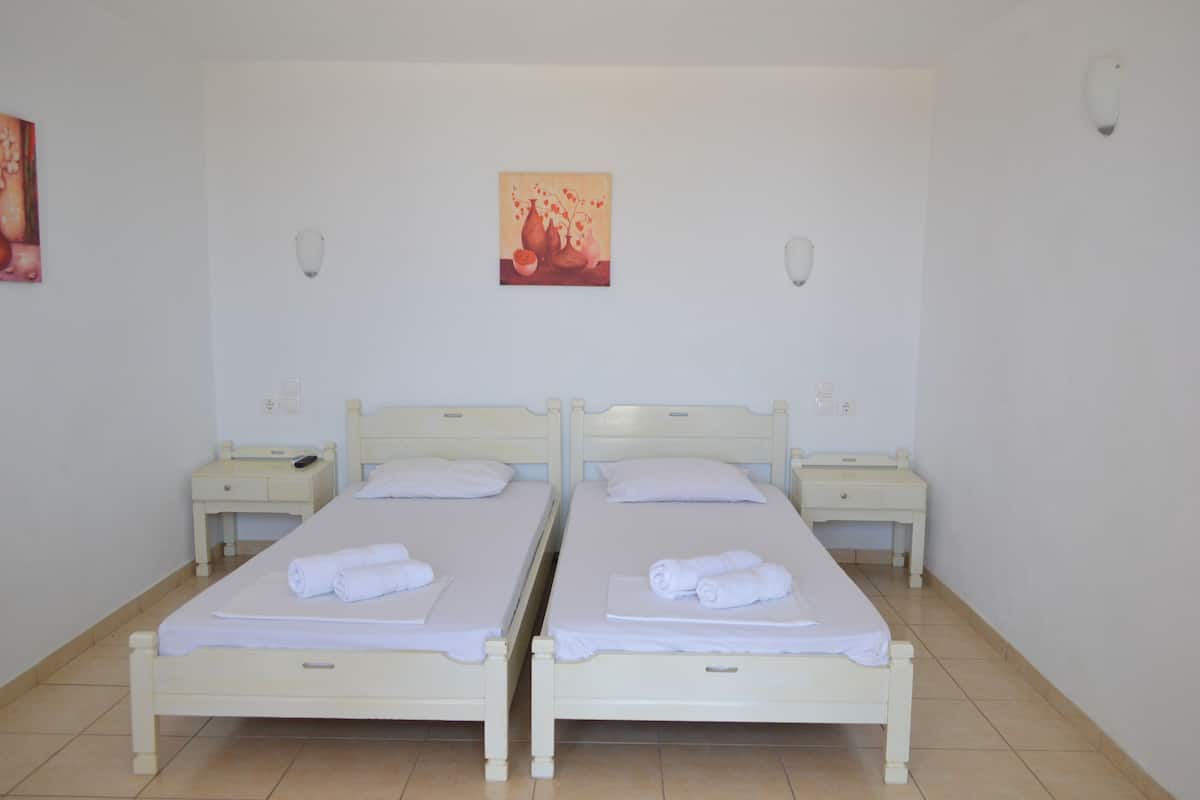 A hotel room with two single beds, each with a white mattress and matching headboard. There are bedside tables with lamps and a painting above the beds.