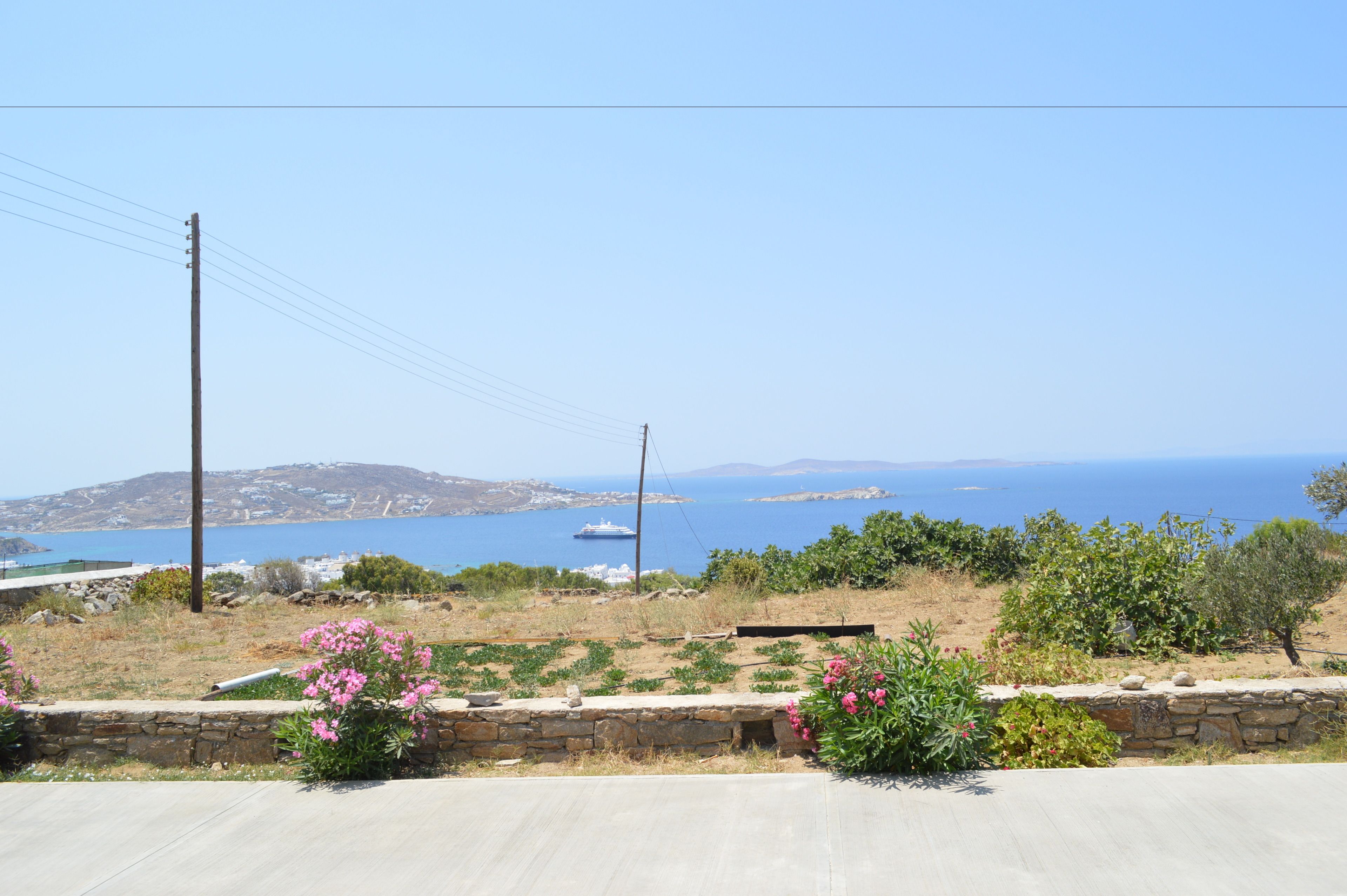Eine Küstenlandschaft mit einer Steinmauer, blühenden Pflanzen und Blick auf das Meer und entfernte Inseln.