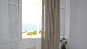 Ein Zimmer mit Meerblick, ein Fenster mit Vorhängen, ein kleiner Tisch mit Blumentopf und ein Holzstuhl.