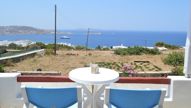 Ein Balkon mit einem weißen Tisch und zwei blauen Stühlen, der auf eine Küstenlandschaft mit freiem Blick auf das Meer und entfernte Inseln blickt.