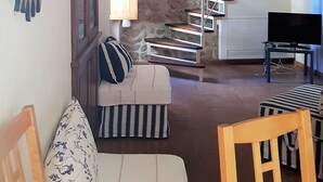 A living room with a sofa, a wooden chair, a striped ottoman, a television, and a staircase against a stone wall.