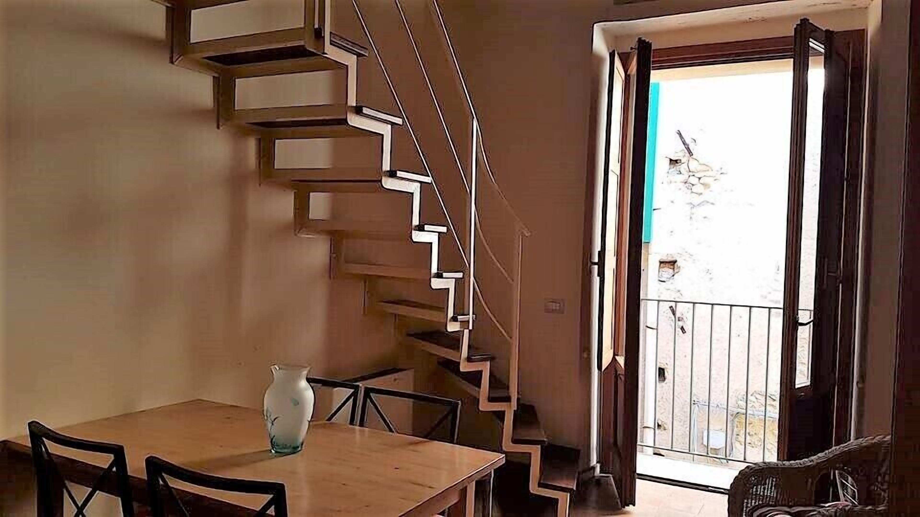 Un coin repas avec une table et des chaises en bois, un vase et un escalier moderne menant à un balcon offrant une vue sur l’extérieur.