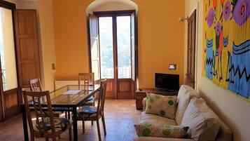Un comedor con una mesa de vidrio y sillas, un sofá con cojines estampados, un televisor sobre una mesita de madera y un cuadro abstracto y colorido en la pared.