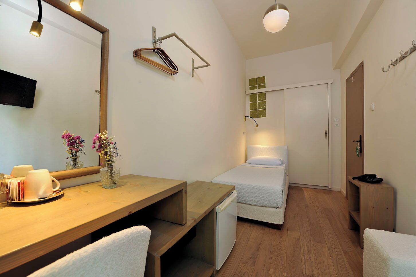 Une chambre d’hôtel compacte comprenant un bureau en bois, un lit avec un matelas blanc, une petite table de chevet, un miroir et un luminaire fixé au mur.
