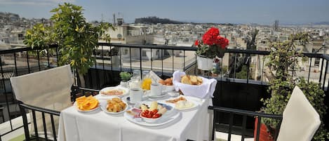A balcony table set with breakfast items, a potted plant, and a view of a cityscape.