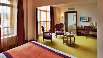 A hotel room with a bed, two chairs, a small table with a vase, a desk with a computer, and a large window with curtains.