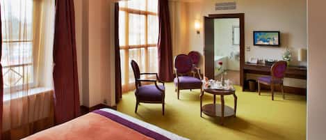 Habitación de hotel con cama, dos sillas, una mesita con un florero, un escritorio con computadora y un ventanal con cortinas.
