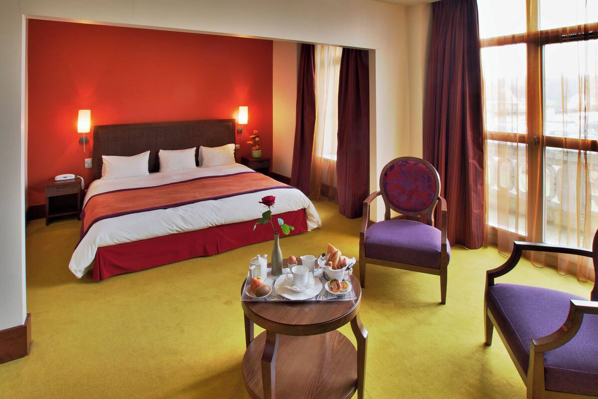 A hotel room with a large bed, a dining table with breakfast items, a chair, and a view of the outside through large windows.