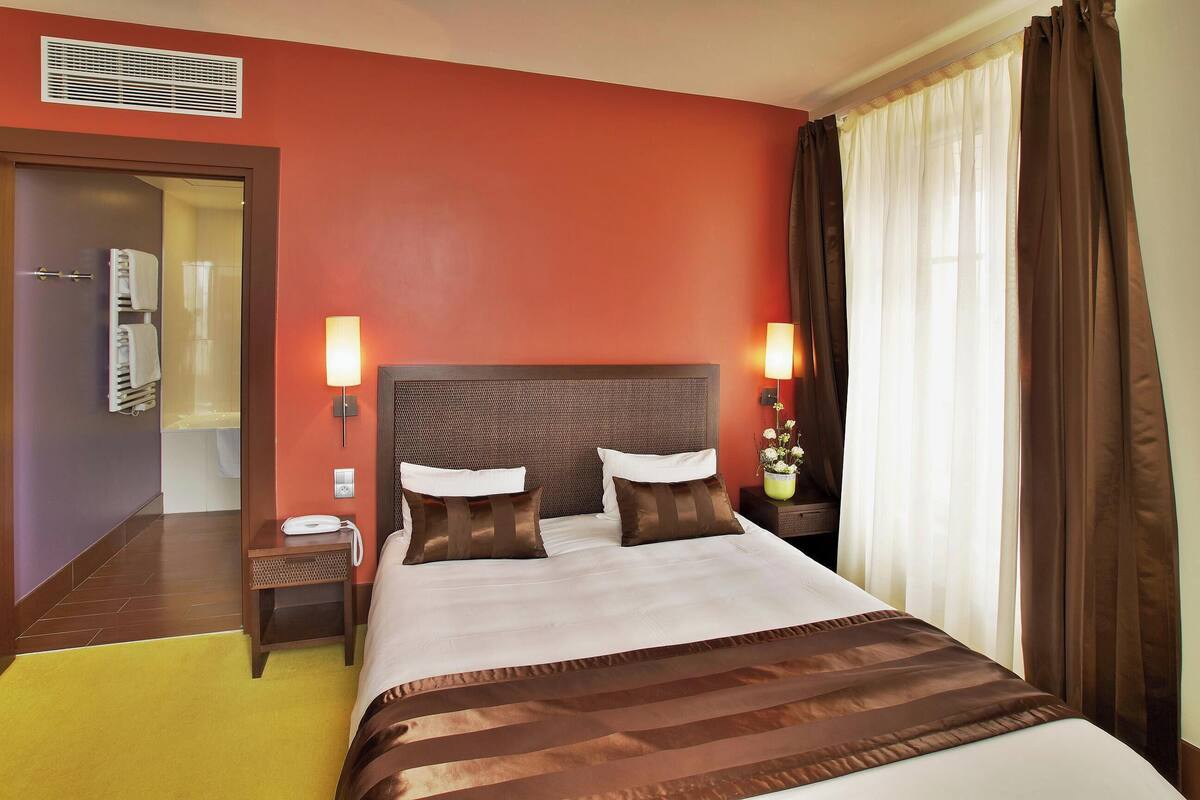A hotel room with a large bed, two bedside lamps, a nightstand, a window with curtains, and a door leading to a bathroom.