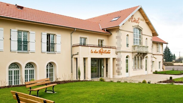 A hotel with a classic architectural style, featuring a central entrance with a glass door, a red-tiled roof, and a well-maintained lawn with benches. - Hotel Le Clos Rebillotte (Luxeuil-les-Bains)