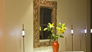 A room with a purple chair, a white ottoman, a table with a vase of yellow flowers, and a large ornate mirror on the wall. - Hotel Le Clos Rebillotte (Luxeuil-les-Bains)