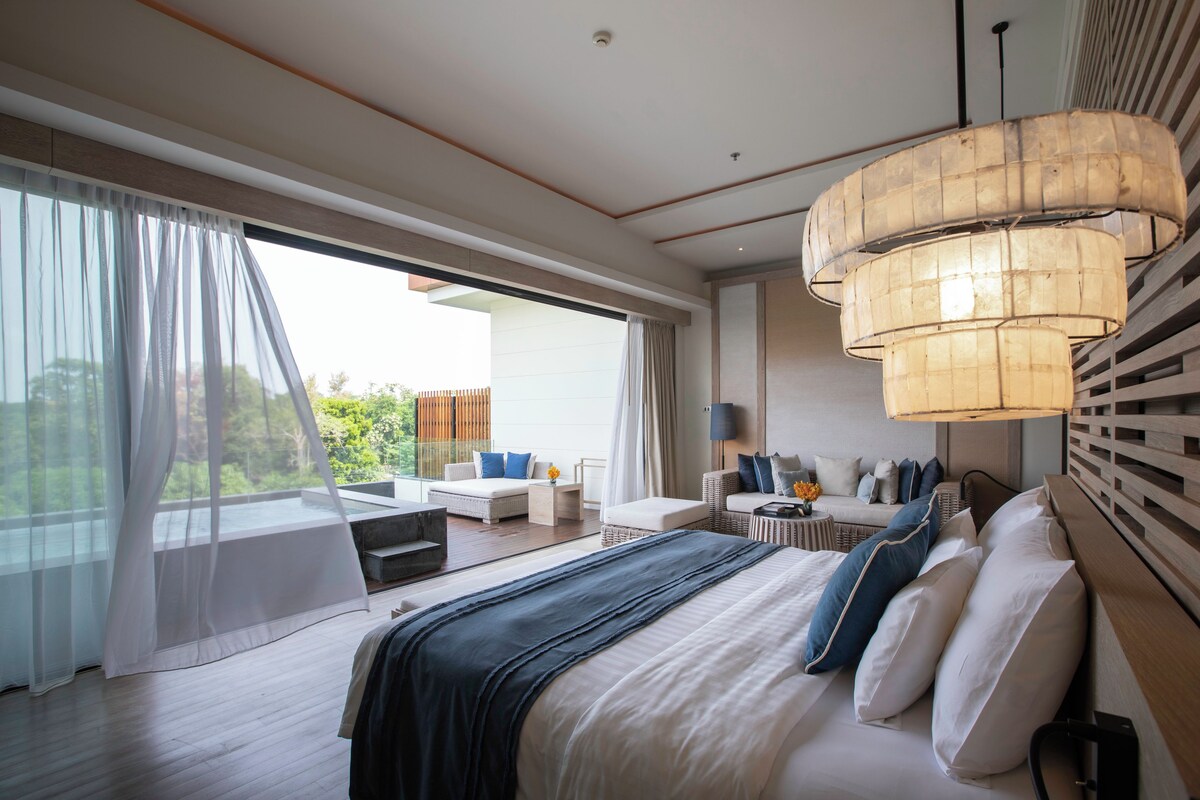 A modern bedroom with a large bed, a seating area with a sofa and chairs, and a large window with sheer curtains.