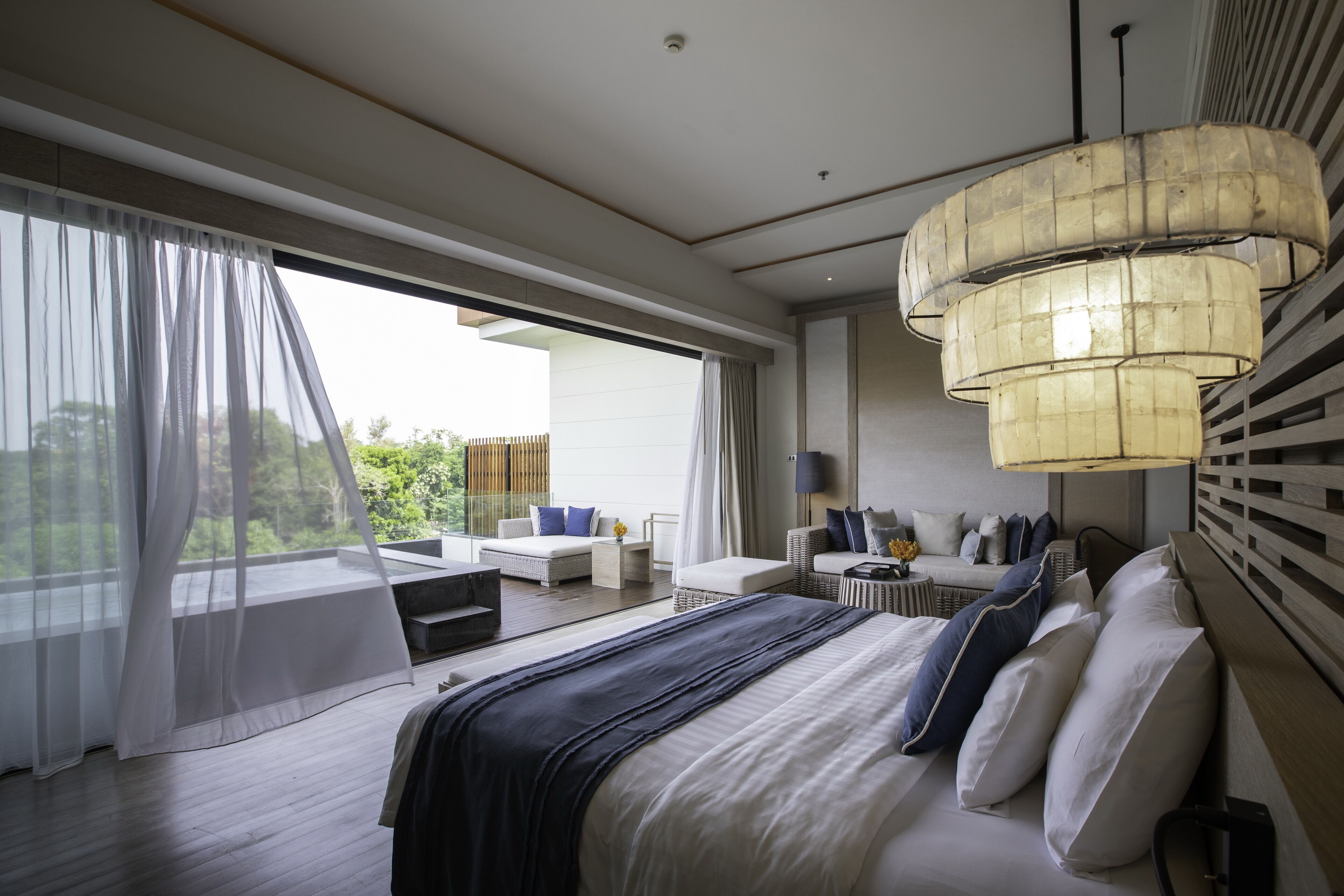 Une chambre moderne avec un grand lit, un coin salon avec un canapé et des fauteuils, et une grande fenêtre ornée de rideaux légers.