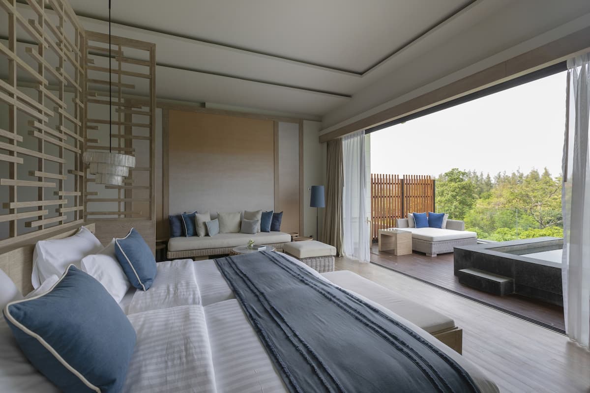 A modern hotel room with a large bed, a sofa, a dining area, and a view of the outdoors.