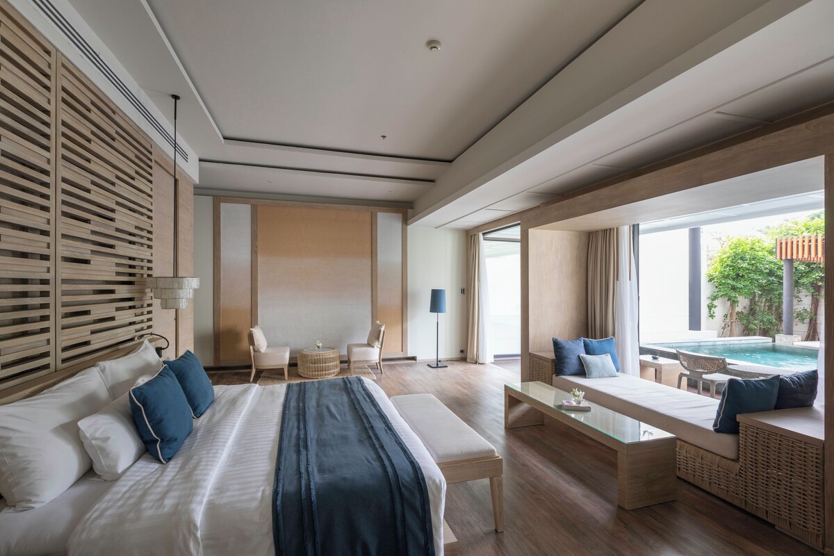 A modern hotel room with a large bed, a seating area with a view of a pool, and a dining table.