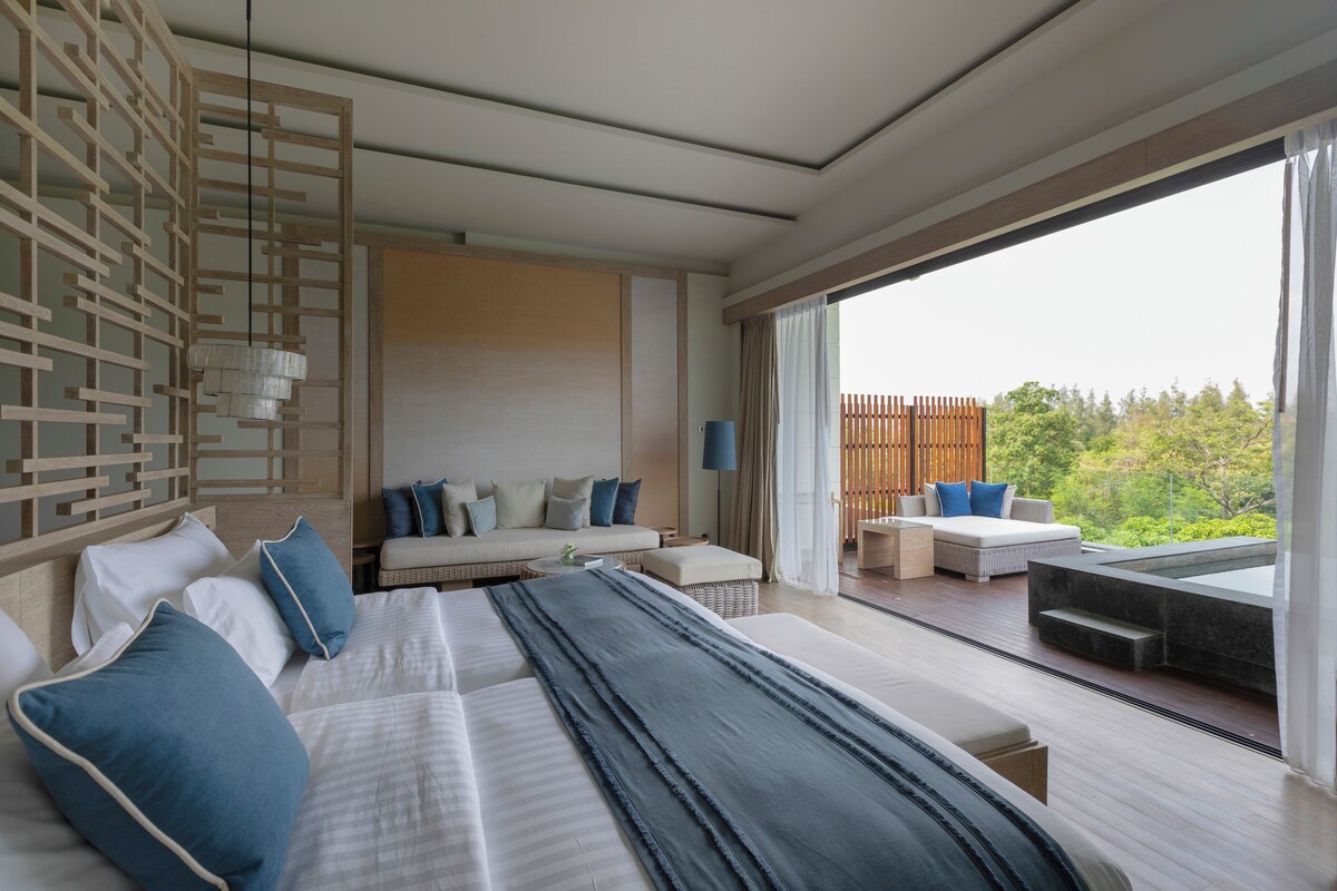 A modern hotel room with a large bed, a sofa, and a balcony with a view of greenery.