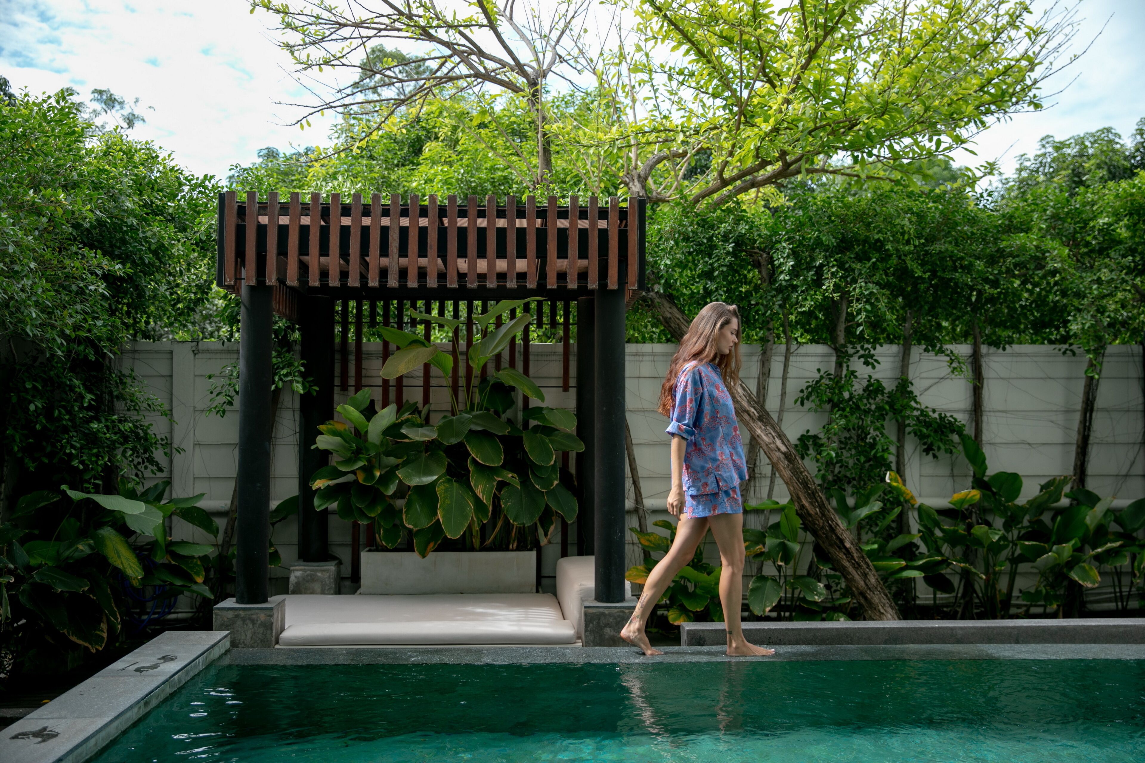 A person walking by a pool with a wooden pergola and lush greenery in the background.