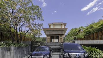 Um edifício moderno de dois andares com telhado plano e entrada central. Há dois carros estacionados na frente, e o edifício é cercado por vegetação e um cercado de madeira.
