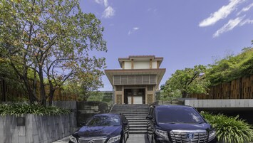 Um edifício moderno de dois andares com telhado plano e entrada central. Há dois carros estacionados na frente, e o edifício é cercado por vegetação e um cercado de madeira.