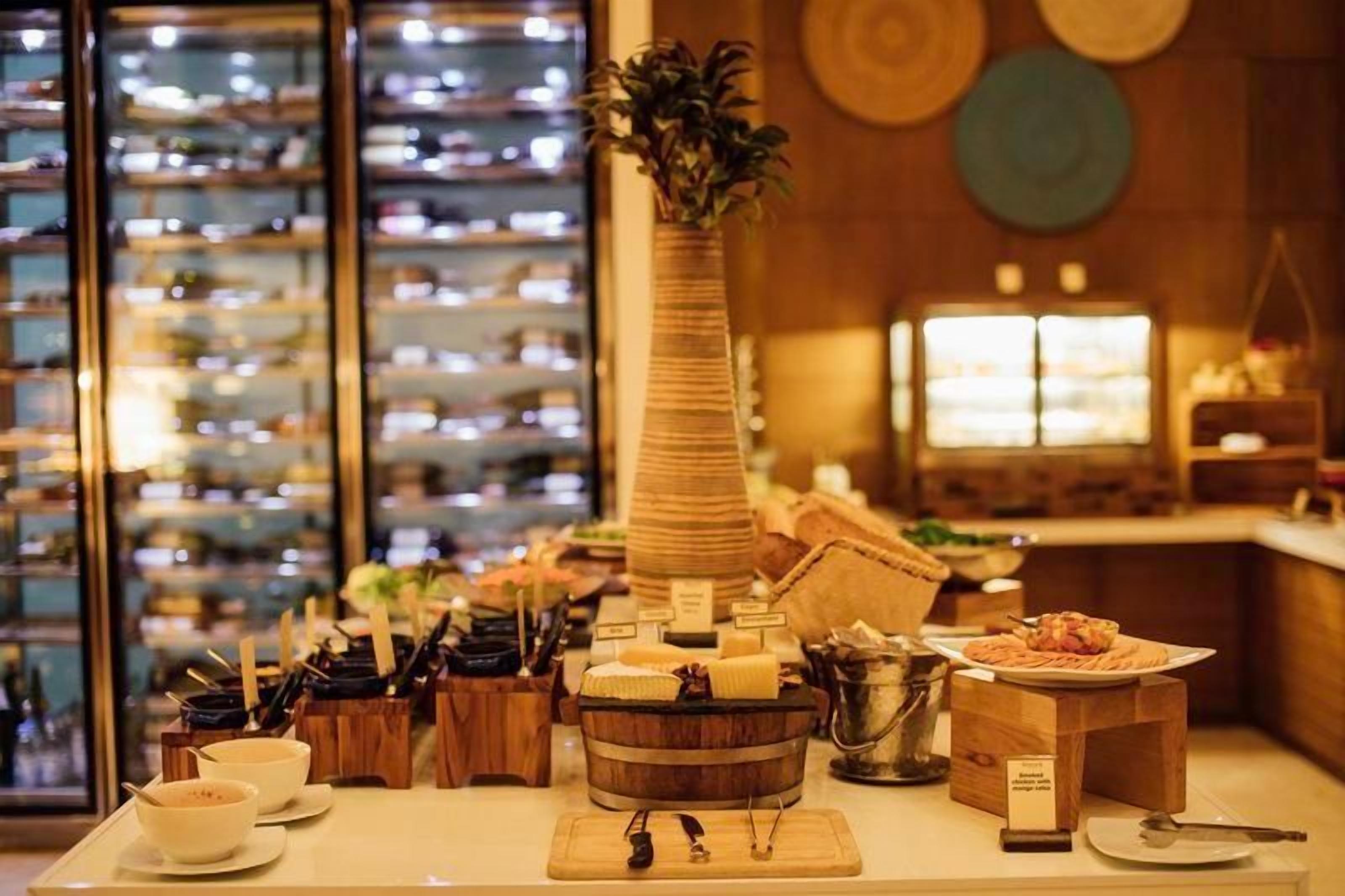 Disposição de buffet com diversos alimentos e utensílios sobre uma mesa, com adega e janela ao fundo.