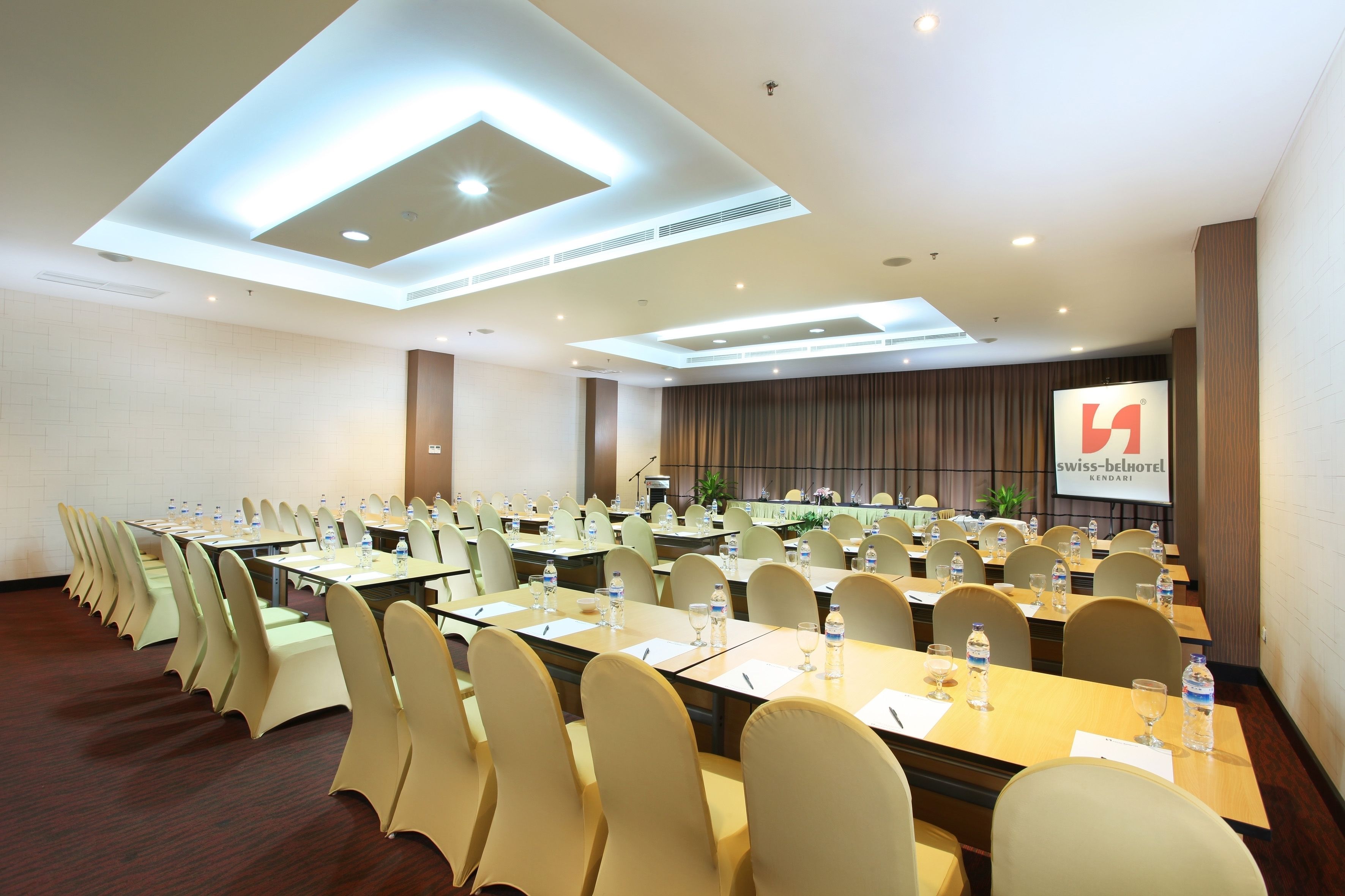 A conference room with multiple tables, chairs, and a large screen displaying the Swiss-Belhotel logo.