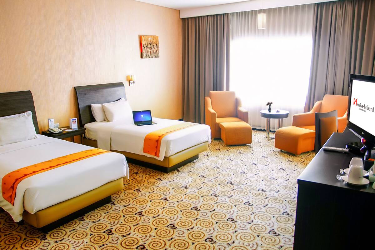 A hotel room with two beds, a desk, a chair, a small table, and a TV.