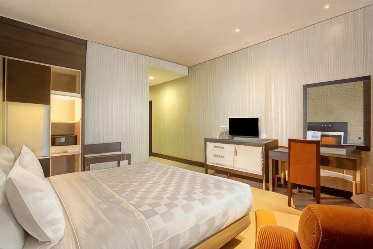 A modern hotel room with a large bed, a desk with a computer, a TV, and a seating area with a chair.