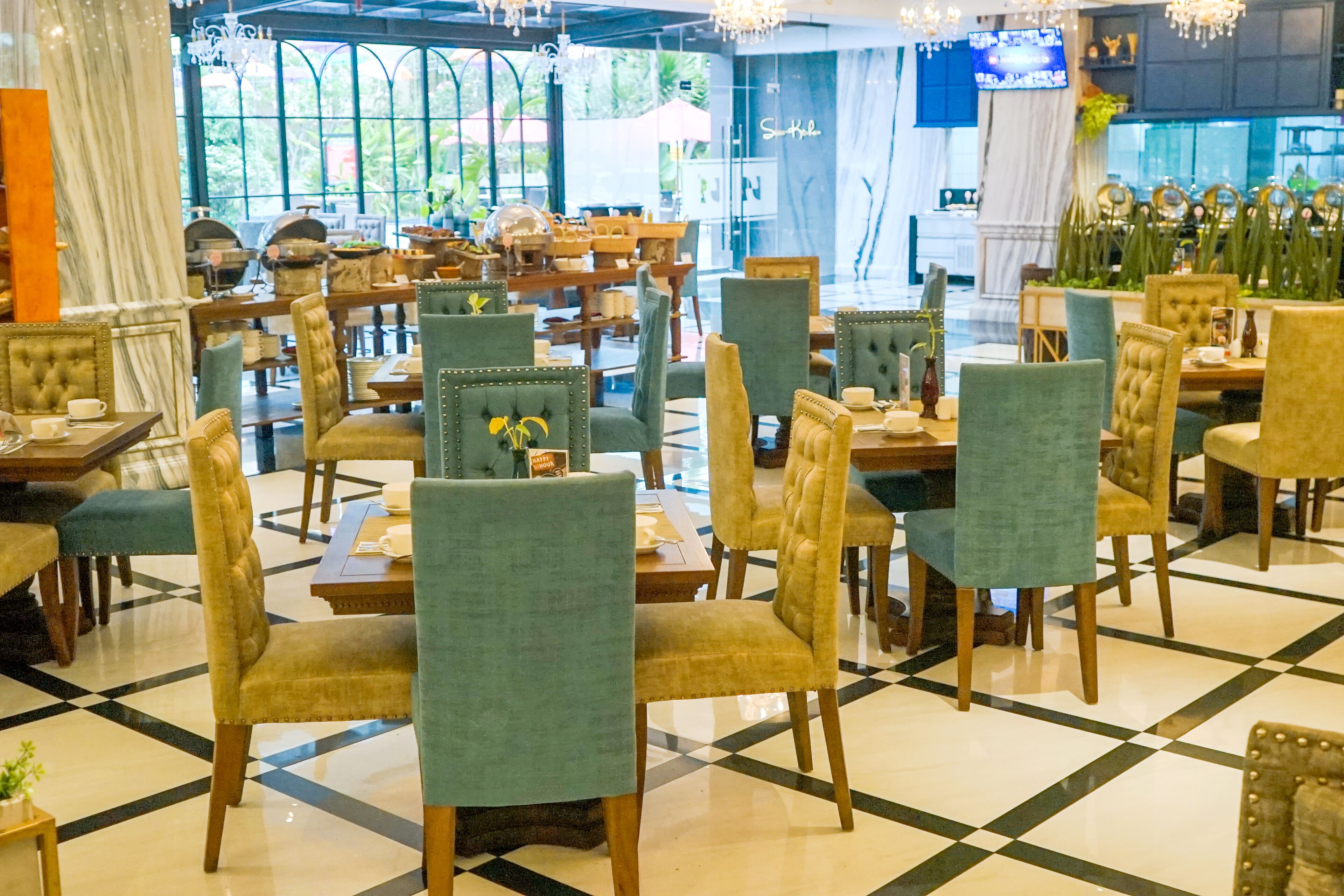 A spacious restaurant with multiple dining tables and chairs, large windows with a view of greenery, and a chandelier hanging from the ceiling.